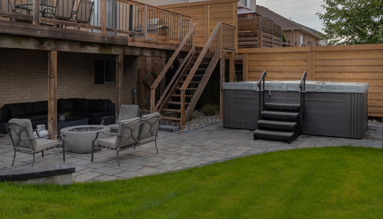 Close up of residential backyard with stone patio, fire pit, green grass, hot tub and privacy fence.