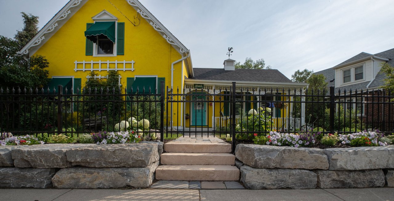 Residential front yard with bright yellow house, colour gardens, stone slabs and black fence